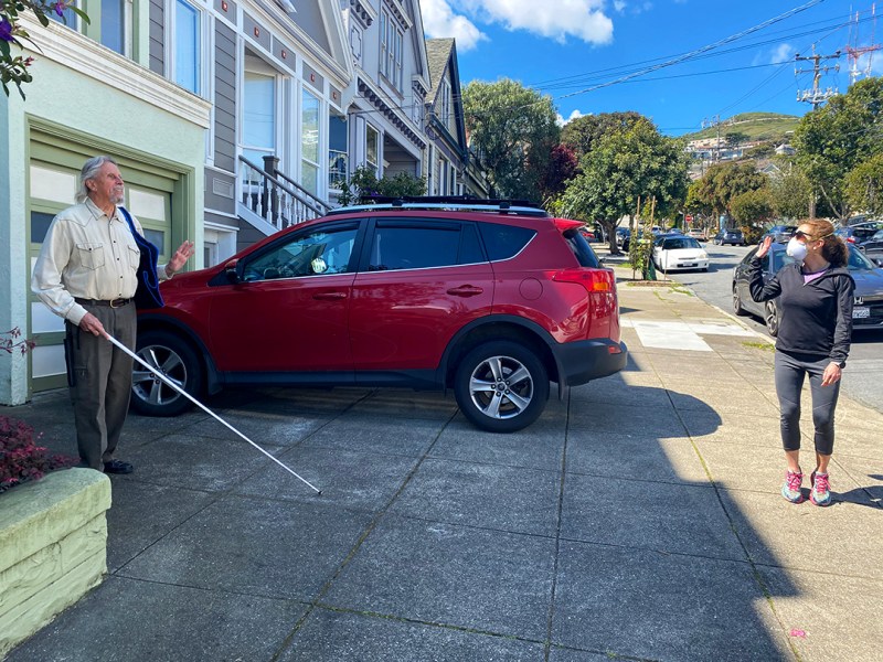 Man holding a white cane waves from a distance to a neighbor standing on the sidewalk and wearing a mask.