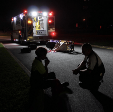 Emergency technician crouches in street at night; emergency vehicle and empty gurney in background