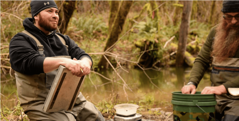 A man looking skyward kneels next to a river, wearing waders and holding a clipboard